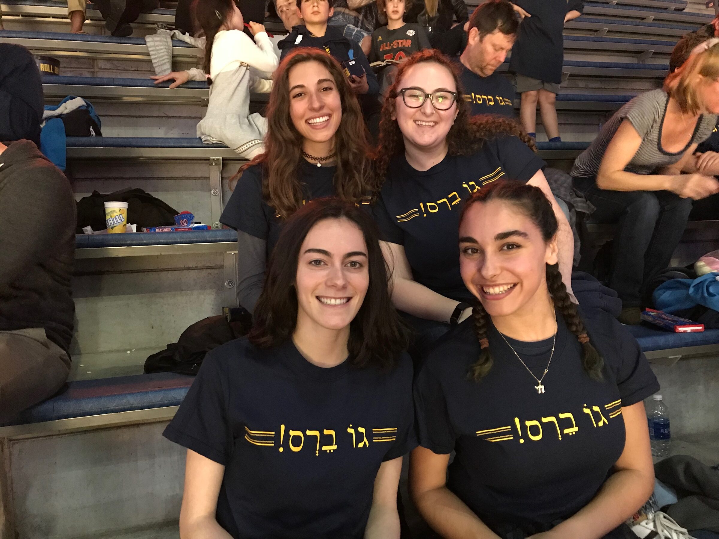 Four students on stadium bleachers wearing shirts reading “Go Bears” in Hebrew